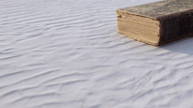 An ancient book lies on a soft, white surface, its pages worn and edges frayed. The gentle texture of the backdrop highlights the books time weathered charm and nostalgic allure.