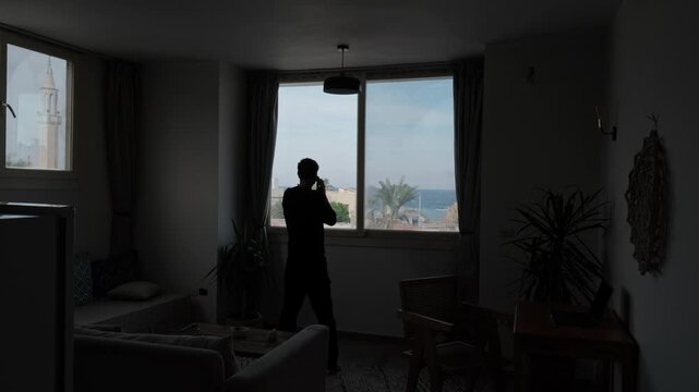 Stressed man having an emotional phone conversation at home, pacing in silhouette by a window overlooking the sea, mountains and palm trees in a dark lifestyle interior during a tense personal moment
