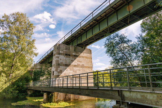 Old steel railway bridge on a stone pier above a river with a footbridge in the foreground, low angle view with green trees and blue sky, historic infrastructure in Germany