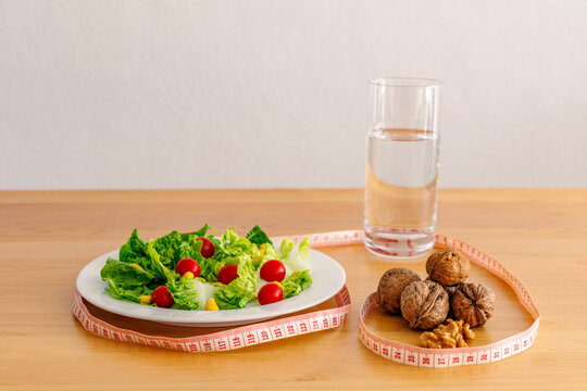 Healthy diet concept with fresh salad plate, walnuts and glass of water surrounded by a measuring tape on wooden table, weight loss, nutrition and healthy eating