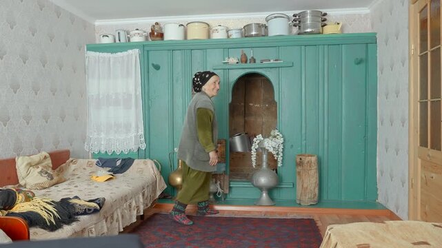Woman standing in profile beside turquoise wooden cabinet inside cozy traditional room, surrounded by lace curtain, patterned wallpaper, flowers, cookware, warm light, muted pastel tones