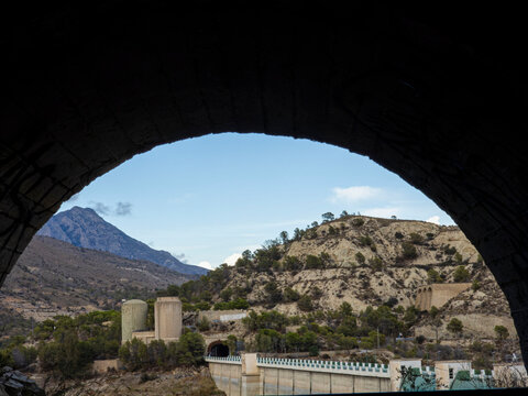 View of arid hills and concrete structures near Amadorio dam framed by dark stone arch