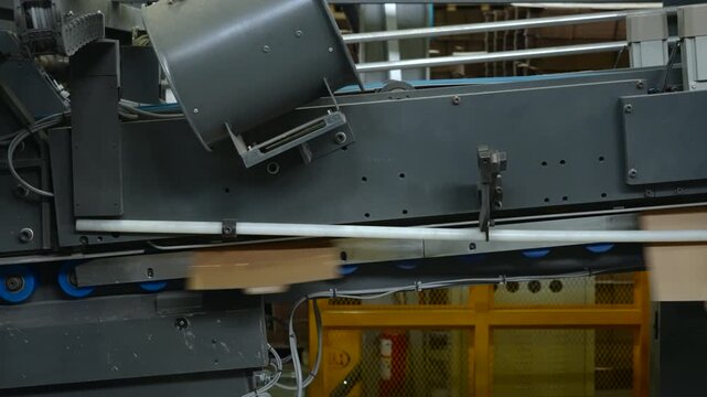 Industrial conveyor machinery moving cardboard boxes inside manufacturing facility, with dark metal panels, rollers, cables, yellow safety fencing, and blurred transport line