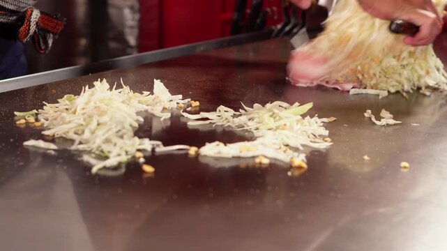 Chef flips Hiroshima okonomiyaki with two metal spatulas on a sizzling teppanyaki griddle and gathers the cabbage. Skillful Japanese food cooking, wide static shot, real time.