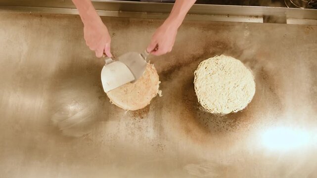 Top view of chef lifting okonomiyaki with two spatulas and stacking it on fried yakisoba noodles on a hot griddle. Hiroshima style layered pancake from above, flat lay.