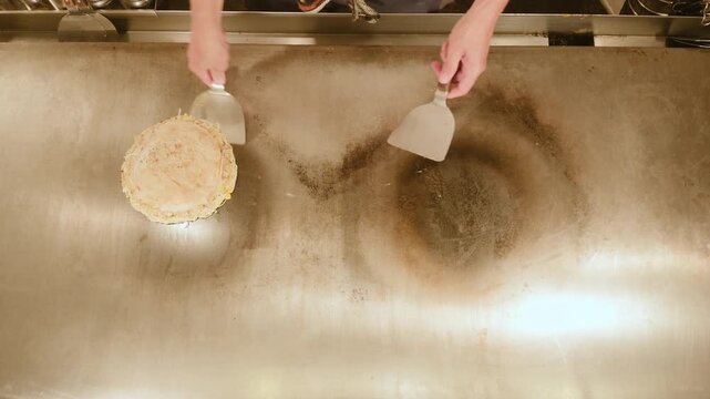 Top view of Hiroshima okonomiyaki placed on a spread egg and flipped with two spatulas on a hot iron griddle, steam rising. Japanese savory pancake from above, flat lay.
