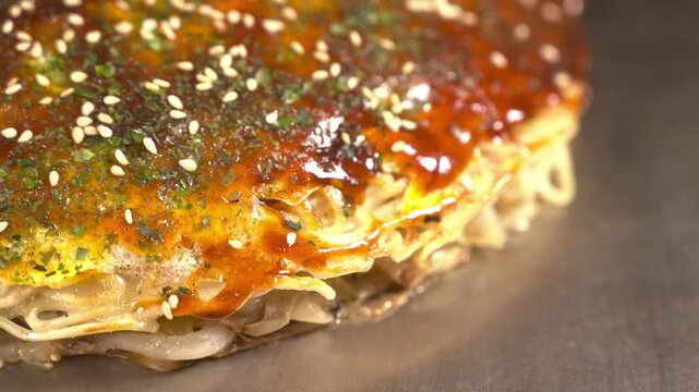 Macro close up of finished Hiroshima okonomiyaki glistening with sauce, aonori and sesame, layers of noodles and egg on a hot griddle. Mouthwatering Japanese food, delicious detail.