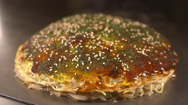 Finished Hiroshima style okonomiyaki with sauce, aonori and sesame on a hot iron griddle in a dark moody teppanyaki restaurant. Delicious Japanese savory pancake, static shot.