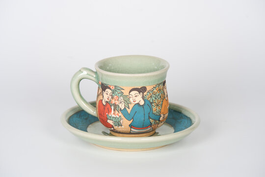 Thai green celadon teacup and saucer painted with traditional villager figures on white background.