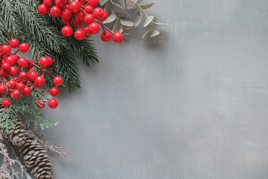 Christmas and New Year frame with spruce twigs, red berries and snow-covered cones on a gray textured background with space for text.