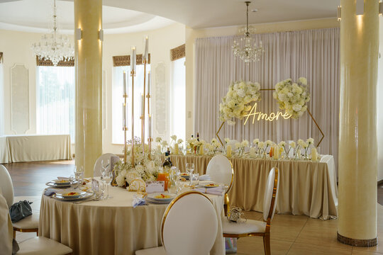 Luxurious wedding reception hall featuring decorated round tables with floral centerpieces, gold candles, and a glowing neon amore sign backdrop