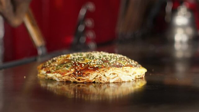 Chef serves Hiroshima okonomiyaki, sliding it across the hot teppanyaki griddle to the customer with spatulas. Japanese restaurant hospitality, warm red interior, static shot.