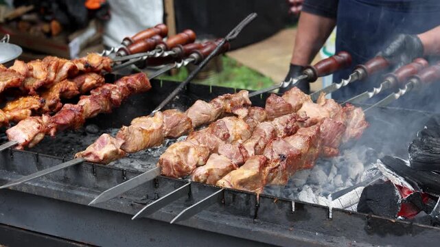Male hand turning metal skewers with shashlik meat pieces cooking over hot charcoal with smoke rising from grill, close up