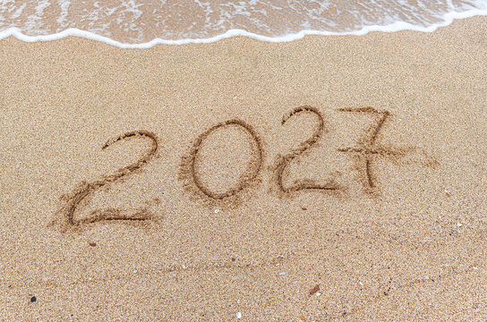 The number 2027 is written in the soft sand near gently washing ocean waves on a sunny beach.