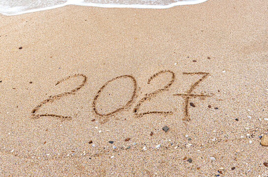 The number 2027 is written in the soft sand near gently washing ocean waves on a sunny beach.