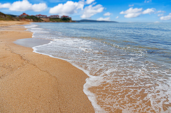 Gentle ocean waves lap against a sandy beach under a bright blue sky with fluffy white clouds.