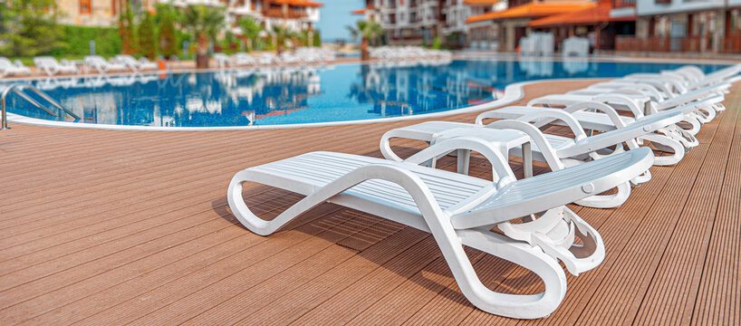 Empty white sun loungers stand in a row on a wooden deck next to a clear blue swimming pool at a tropical resort.