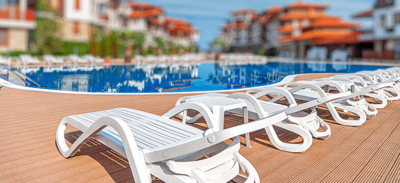 A row of white sun loungers sits beside a bright blue swimming pool under a sunny blue sky.