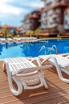 Empty sun loungers sit on a wooden deck by a bright blue swimming pool.