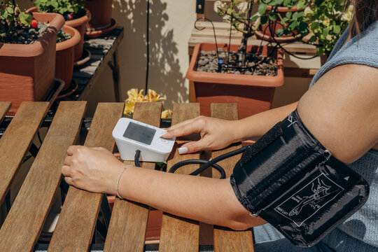 Woman Checking Blood Pressure With Digital Monitor On Balcony Table Surrounded By Potted Plants