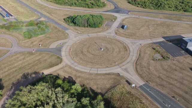 High altitude drone perspective looking down at a specialized automotive testing facility with a paved circular intersection and surrounding dry grass terrain during daylight.