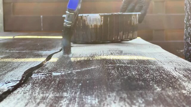 Low angle close-up of a paintbrush applying dark wood protective stain to a timber board. Ideal for DIY tutorials, wood preservation product ads, and carpentry media.