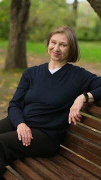 Vertical video of beautiful smiling mature blond woman sitting on the bench in park