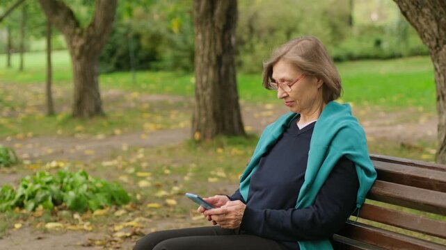 Beautiful smiling mature blond woman with glasses using her phone for texting messages while sitting on bench in the park