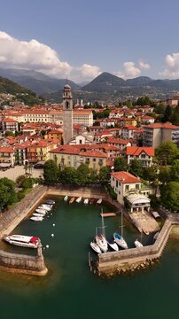 Drone flyover of Pallanza harbor in Verbania, Lake Maggiore, with boats and sailing yachts, historic architecture, bell tower, red tiled roofs and the town stretching into the background.
