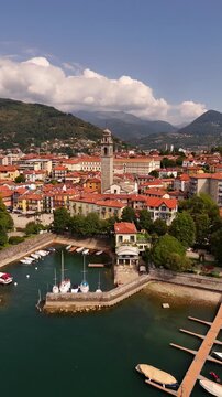 Drone flyover of Pallanza harbor in Verbania, Lake Maggiore, with boats and sailing yachts, historic architecture, bell tower, red tiled roofs and the town stretching into the background.