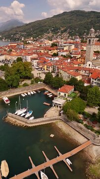 Drone flyover of Pallanza harbor in Verbania, Lake Maggiore, with boats and sailing yachts, historic architecture, bell tower, red tiled roofs and the town stretching into the background.