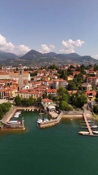 Drone flyover of Pallanza harbor in Verbania, Lake Maggiore, with boats and sailing yachts, historic architecture, bell tower, red tiled roofs and the town stretching into the background.