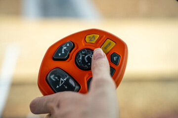 A hand presses a button on an orange handheld controller with a joystick against a blurred indoor background. © Marina