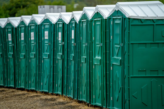 A row of green portable toilet cabins with white roofs stands on bare ground outdoors, with trees in the background.