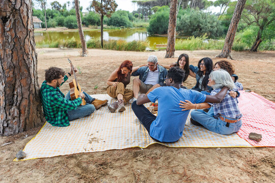 Group sitting on a picnic blanket by a pond while a man plays guitar