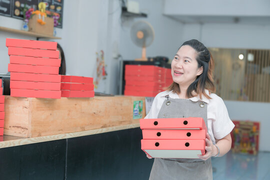 Smiling Asian female pizzeria owner carrying red pizza boxes in restaurant