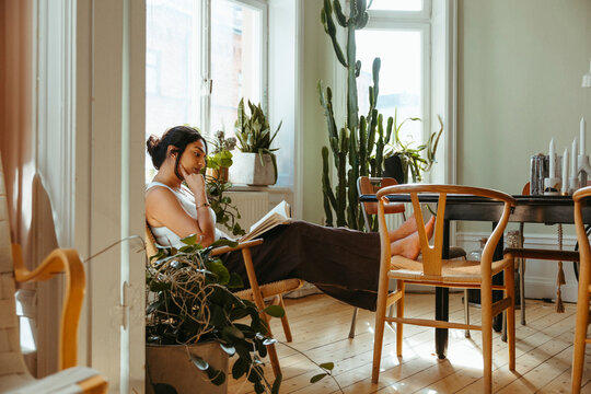 Carefree woman reading book while sitting on chair with feet up in home