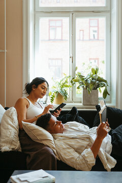 Young man reading book and lying on sofa near girlfriend using smart phone in living room at home