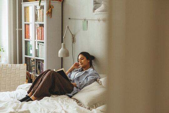Full length of woman spending leisure time reading book while sitting on bed in home