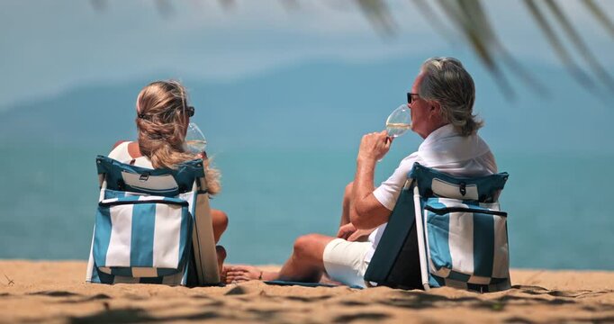 Mature couple on tropical beach in Thailand, enjoy their best life, seated on resort chairs, toasting wine glasses, enjoying luxury, carefree retirement vacation by the sea on Samui island. Back view