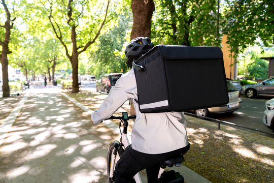 Rear view of delivery man carrying thermal parcel box riding cycle on road