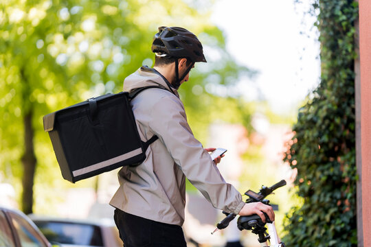 Young delivery man carrying parcel bag standing with bicycle while using smart phone at street