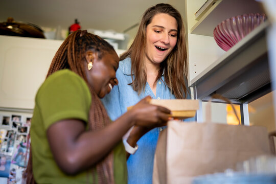 Woman giving facial expression while standing near friend taking out parcel from paper bag at home