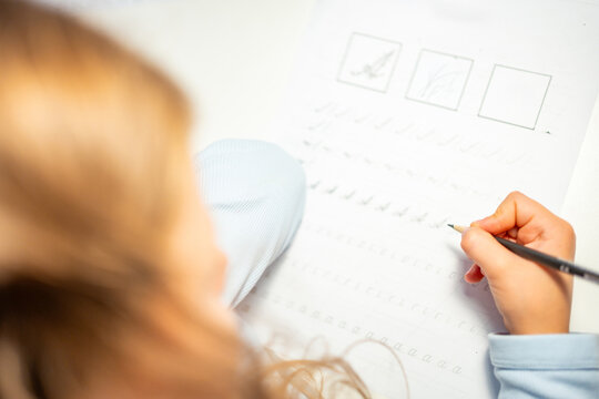 First grade girl practices handwriting with pencil in school workbook. Child learns to write letters during early education with selective focus and shallow depth of field