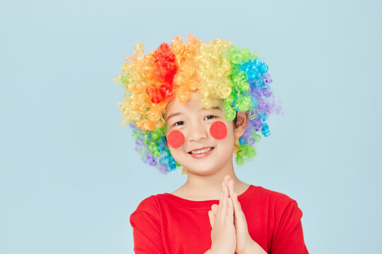 Smiling child in rainbow clown wig and red nose with hands clasped
