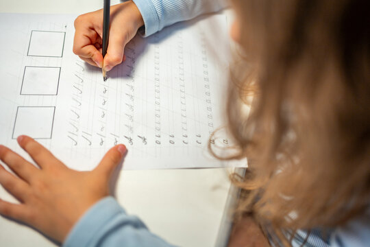 First grade girl practices handwriting with pencil in school workbook. Child learns to write letters during early education with selective focus and shallow depth of field