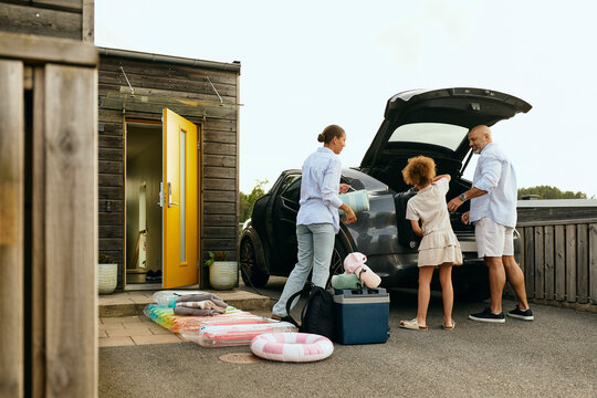 Girl helping parents in loading luggage in electric car for vacation trip at backyard under clear sky