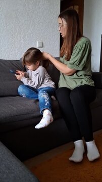 Mother styles daughter's hair while she plays with phone on couch in