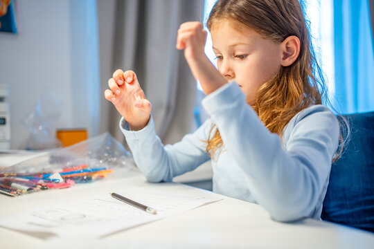 First grade girl stretches tired fingers during handwriting practice at home. Child takes a break from writing to relieve hand fatigue with selective focus