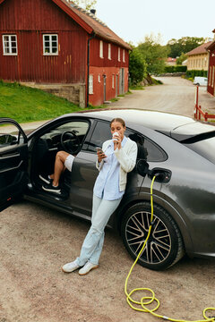 High angle view of woman sipping drink and using smart phone while car getting charged at outdoor charging station
